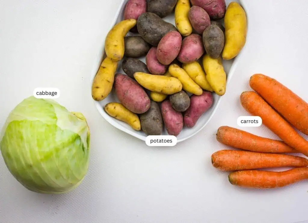A head of cabbage, a plate of assorted fingerling potatoes, and four carrots are arranged on a white surface, each labeled.