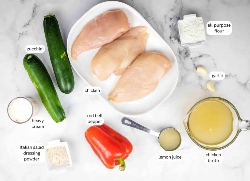 Ingredients for a chicken dish arranged on a countertop, including chicken breasts, zucchini, red bell pepper, heavy cream, broth, flour, garlic, lemon juice, and Italian dressing powder.