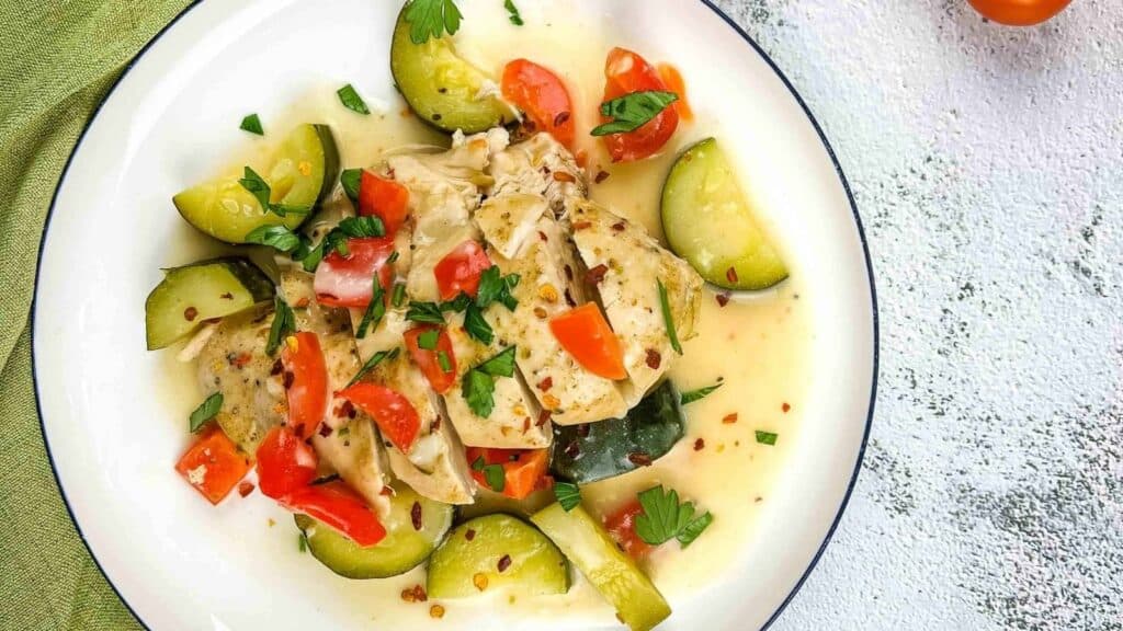 A plate of cooked chicken with zucchini and red bell pepper pieces in a light sauce, garnished with chopped herbs.