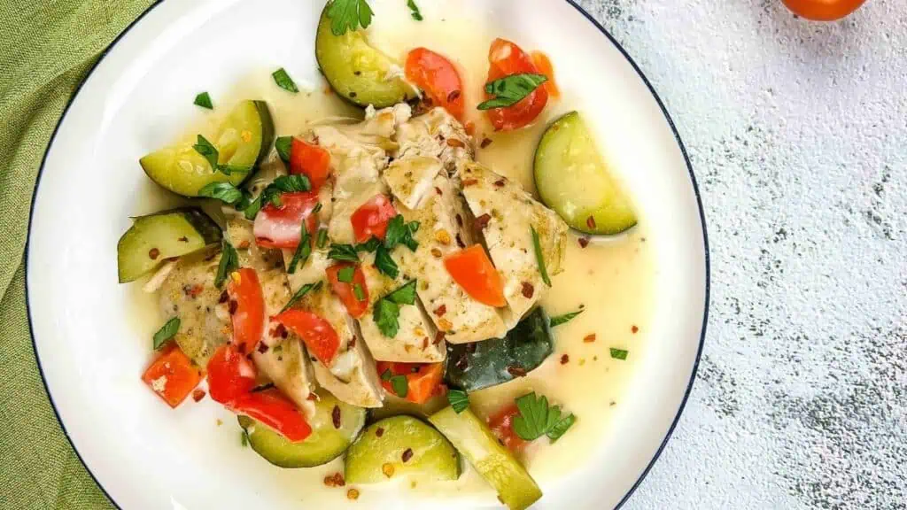 A plate of cooked chicken with zucchini and red bell pepper pieces in a light sauce, garnished with chopped herbs.