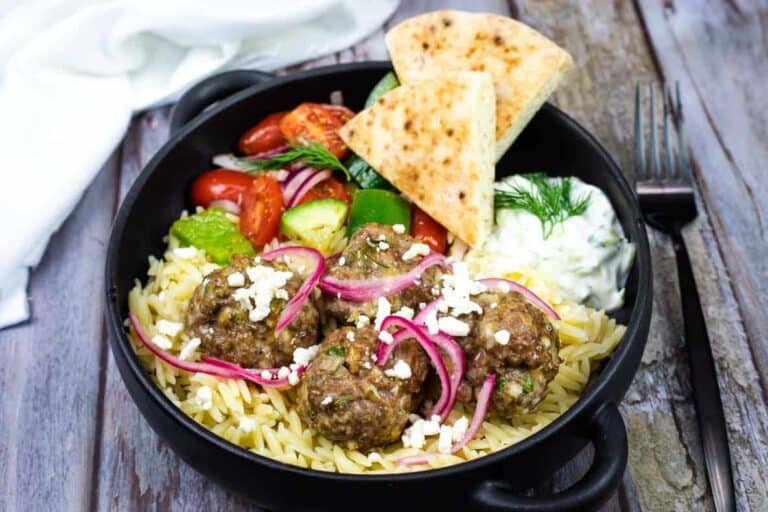 A black bowl with orzo pasta, meatballs topped with feta and red onion, Greek salad, pita bread, and a side of tzatziki sauce.