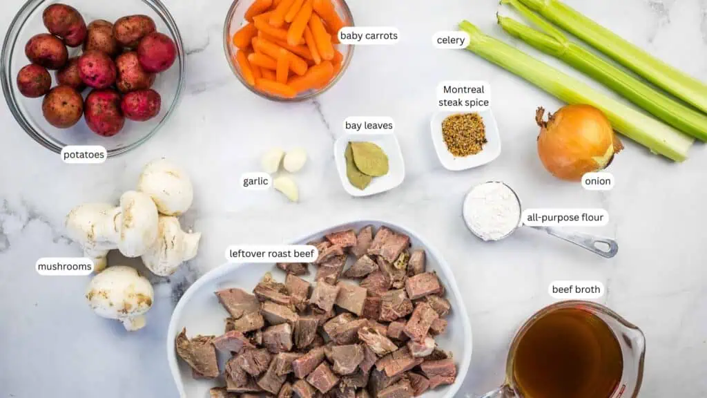 Ingredients for beef stew displayed on a countertop, including potatoes, baby carrots, celery, mushrooms, garlic, onion, bay leaves, Montreal steak spice, flour, roast beef, and beef broth.