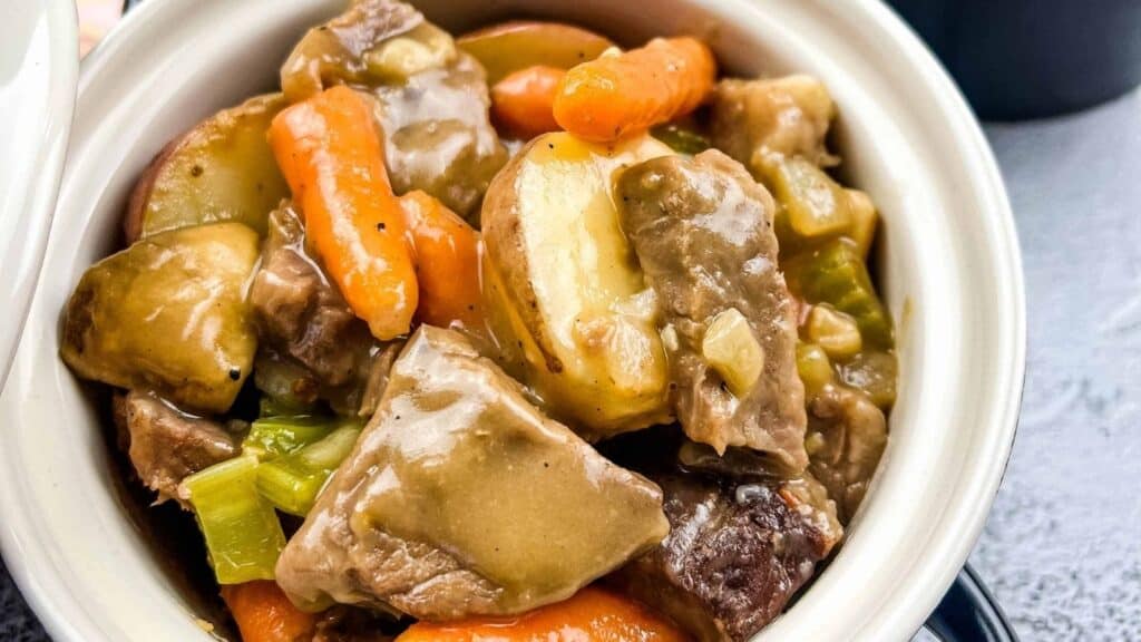 A close-up of a bowl filled with beef stew containing chunks of beef, potatoes, carrots, celery, and a thick brown gravy.