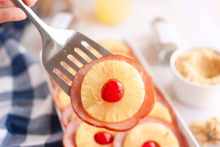A metal spatula holding a slice of ham topped with a pineapple ring and a maraschino cherry; similar slices are visible in the background.