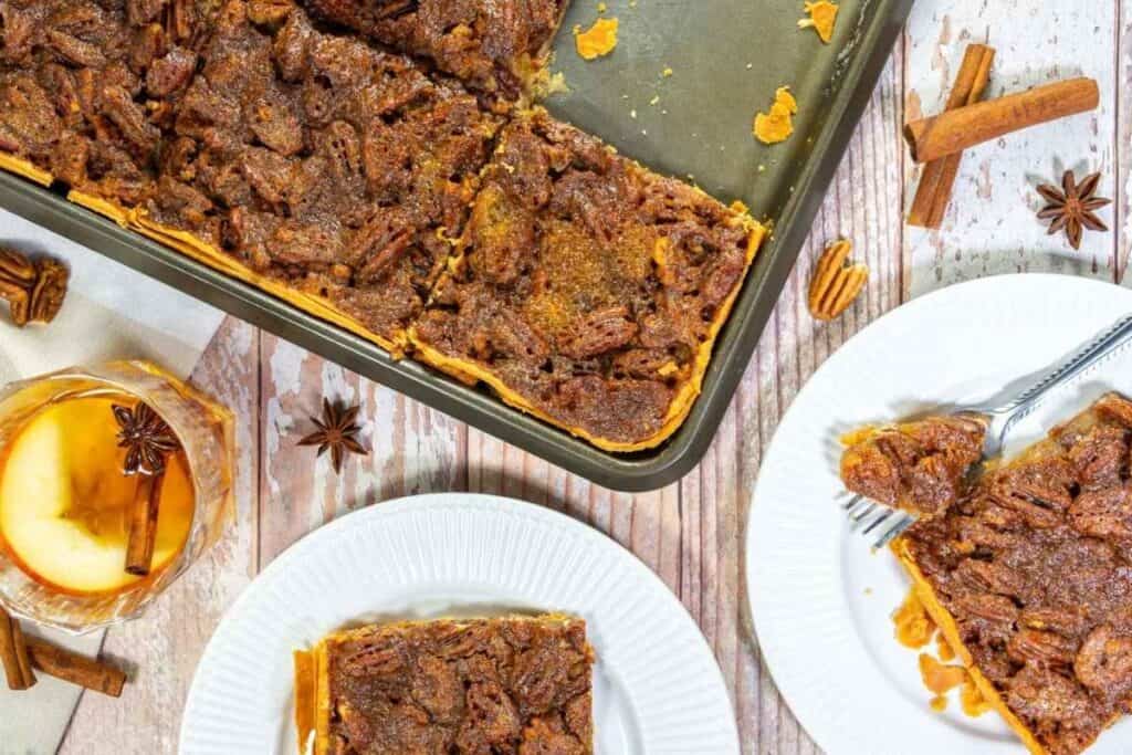 A baking pan with pecan pie bars, two white plates with bars on them, a glass of apple cider, and cinnamon sticks on a wooden table.