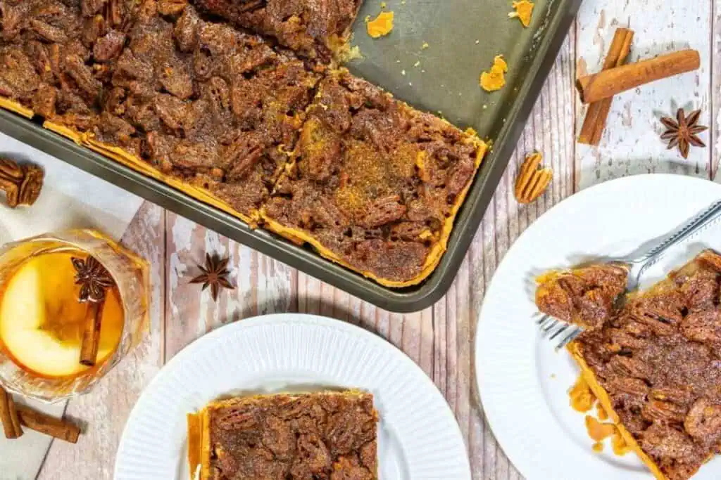 A baking pan with pecan pie bars, two white plates with bars on them, a glass of apple cider, and cinnamon sticks on a wooden table.