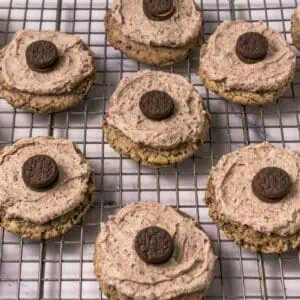 Chocolate chip cookies topped with swirled chocolate frosting and a mini chocolate sandwich cookie, arranged on a cooling rack.