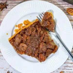 A slice of pecan pie with a fork on a white plate, surrounded by spices and nuts on a wooden surface.