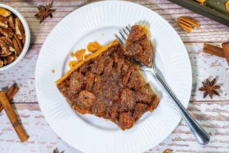 A slice of pecan pie with a fork on a white plate, surrounded by spices and nuts on a wooden surface.