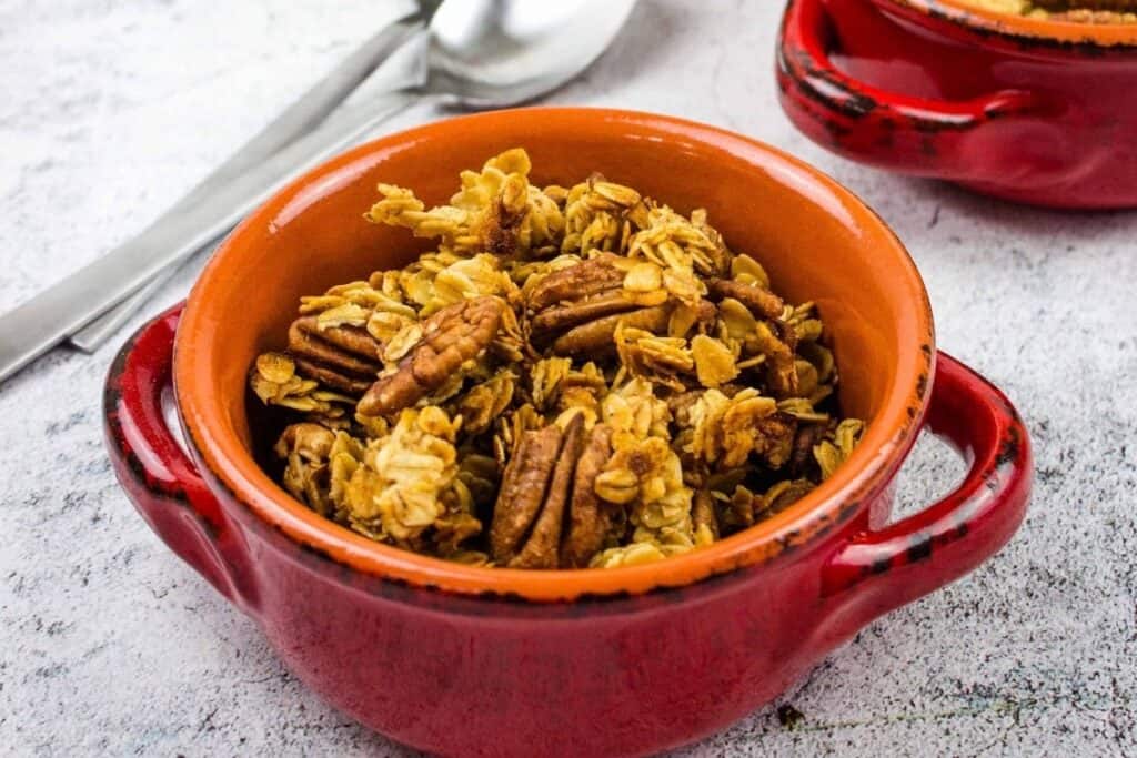 A red bowl filled with granola and pecans sits on a light surface, with a spoon and another bowl in the background.