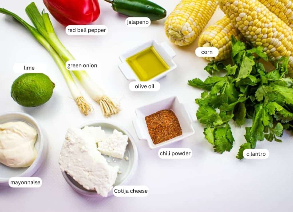Assorted ingredients for Mexican street corn, including corn, cilantro, Cotija cheese, mayonnaise, lime, green onion, red bell pepper, jalapeno, olive oil, and chili powder, on a white surface.