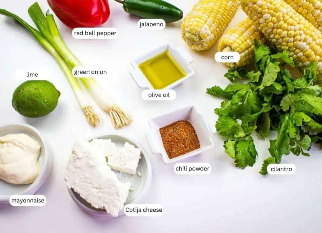 Assorted ingredients for Mexican street corn, including corn, cilantro, Cotija cheese, mayonnaise, lime, green onion, red bell pepper, jalapeno, olive oil, and chili powder, on a white surface.