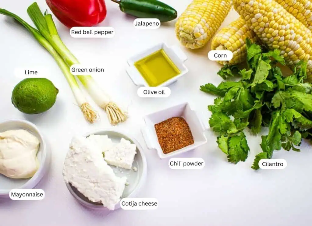Assorted ingredients for Mexican street corn salad with labels on a white surface, including corn, cilantro, chili powder, olive oil, cotija cheese, mayonnaise, lime, green onion, red bell pepper, and jalapeno.