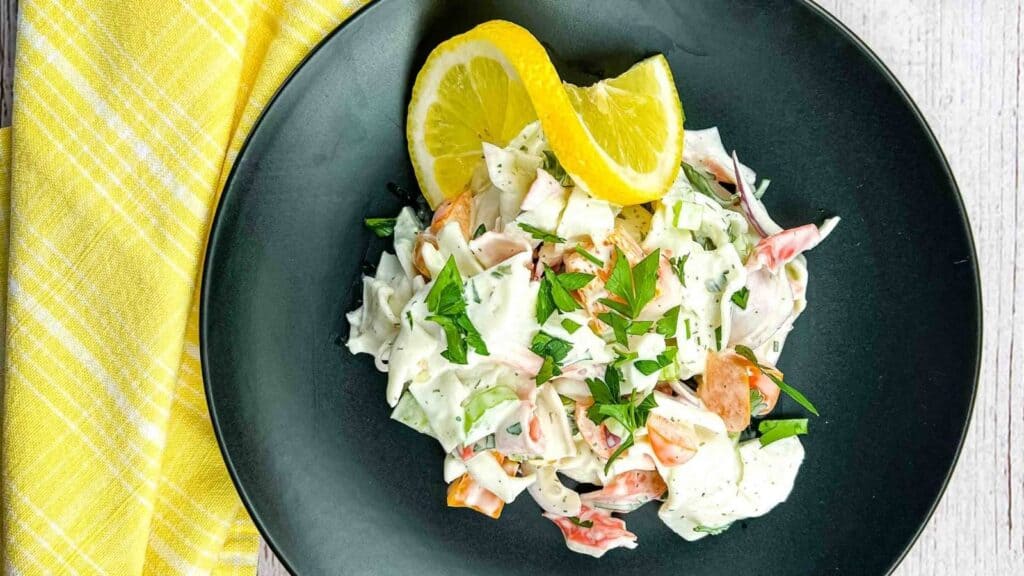 A black plate with a creamy seafood salad garnished with chopped herbs and two lemon wedges, next to a yellow checkered napkin on a light wooden surface.