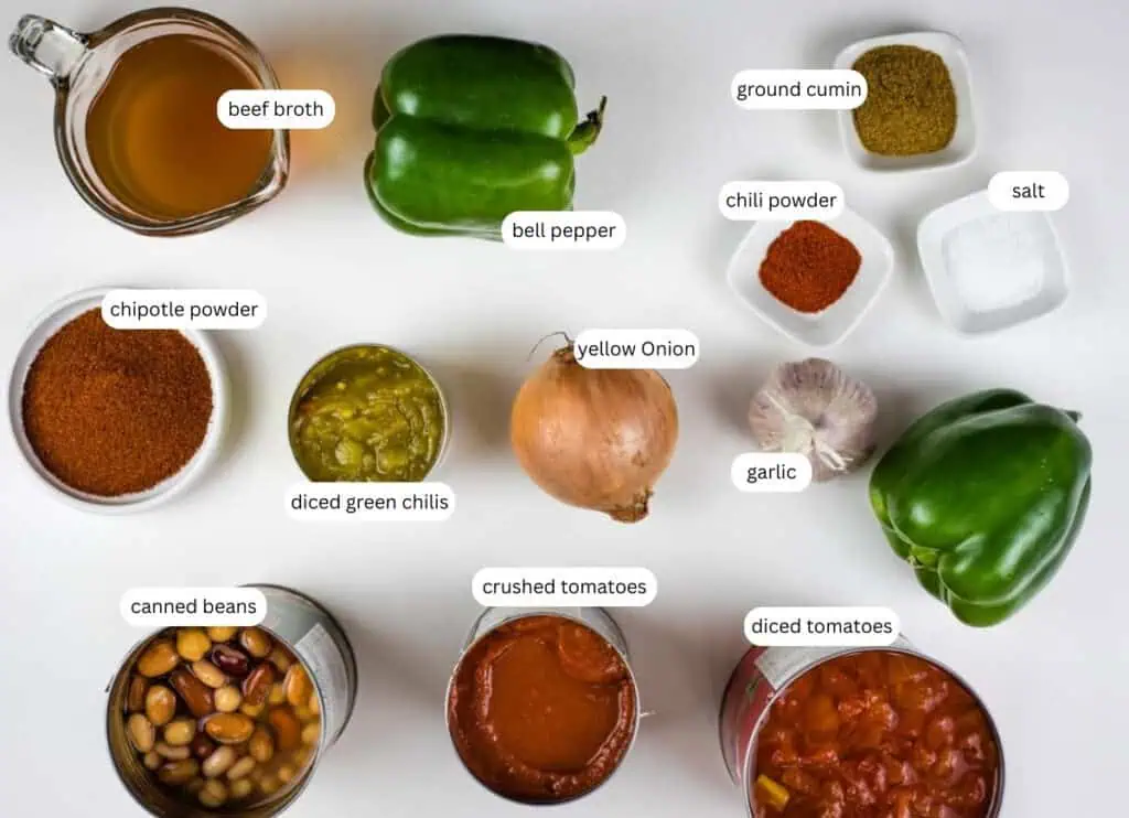 Top-down view of chili ingredients, including beef broth, bell peppers, ground cumin, chili powder, salt, chipotle powder, diced green chilis, yellow onion, garlic, canned beans, crushed tomatoes, and diced tomatoes.