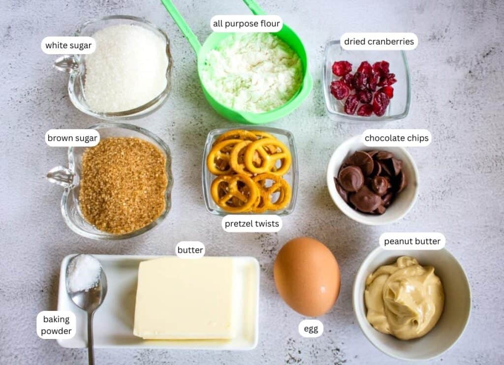 Various baking ingredients are arranged on a table, including white and brown sugar, flour, dried cranberries, chocolate chips, pretzel twists, baking powder, butter, an egg, and peanut butter.