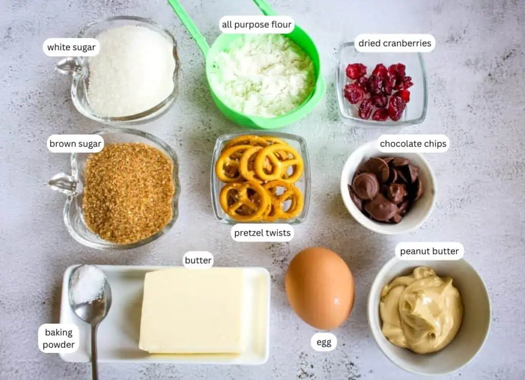 Various baking ingredients are arranged on a table, including white and brown sugar, flour, dried cranberries, chocolate chips, pretzel twists, baking powder, butter, an egg, and peanut butter.