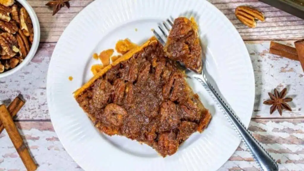 A square slice of pecan pie bar on a white plate with a fork, surrounded by pecans, cinnamon sticks, and star anise on a rustic wooden surface.