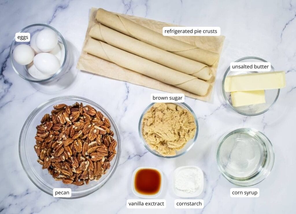 Ingredients for pecan pie are arranged on a marble surface, including eggs, pecans, pie crusts, butter, brown sugar, corn syrup, vanilla extract, and cornstarch.