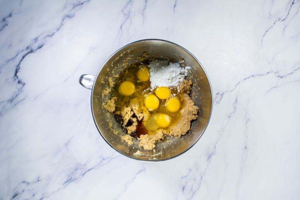 A metal mixing bowl with cookie dough, six cracked eggs, flour, and other ingredients on a white marble surface.