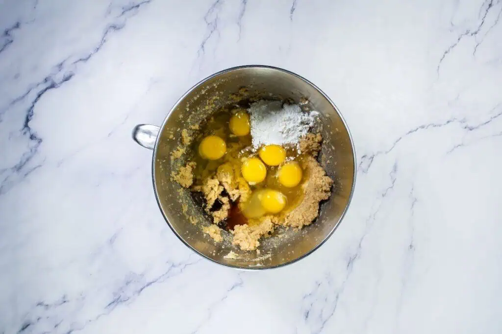 A metal mixing bowl with cookie dough, six cracked eggs, flour, and other ingredients on a white marble surface.