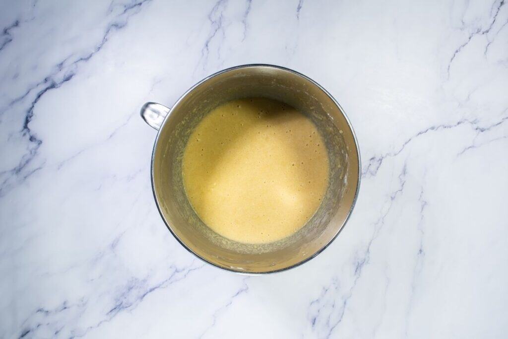 A metal mixing bowl filled with a smooth, light brown batter sits on a white marble surface.
