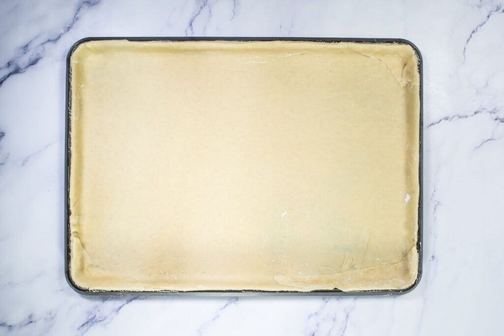 A rectangular metal baking pan filled with raw, evenly rolled dough, resting on a white marble surface.