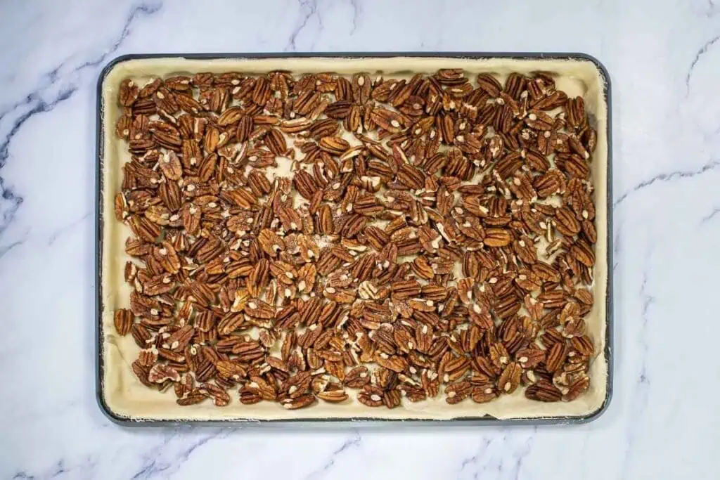A baking sheet lined with unbaked pie crust dough topped with an even layer of raw pecan halves on a marble surface.