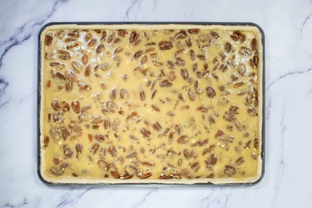 A rectangular baking tray filled with unbaked pecan pie bars mixture on a marble surface.