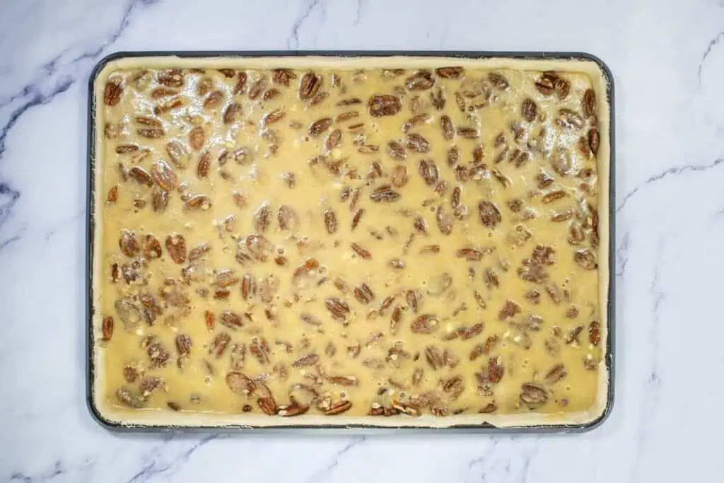 A rectangular baking tray filled with unbaked pecan pie bars mixture on a marble surface.