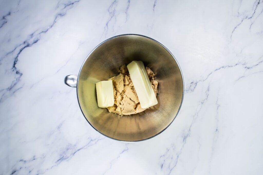 A metal mixing bowl containing two sticks of butter and a mound of brown sugar on a white marble surface.