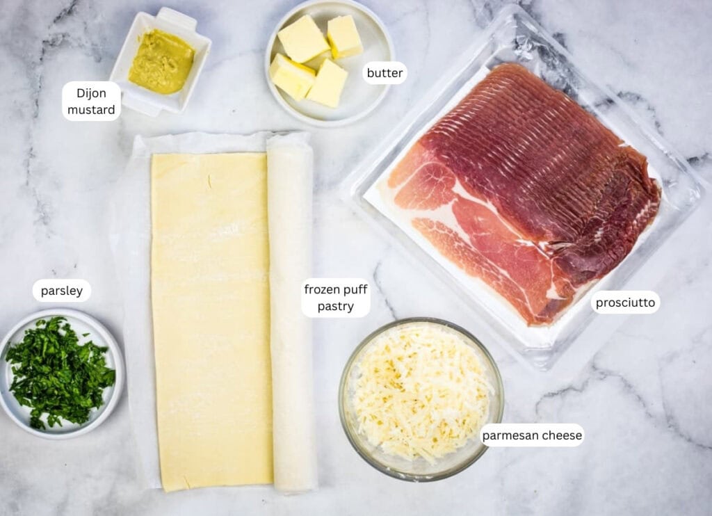 Ingredients displayed on a countertop: Dijon mustard, butter, prosciutto, frozen puff pastry, chopped parsley, and grated parmesan cheese.