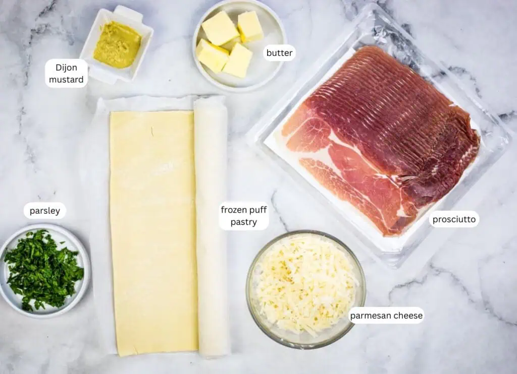 Ingredients displayed on a countertop: Dijon mustard, butter, prosciutto, frozen puff pastry, chopped parsley, and grated parmesan cheese.