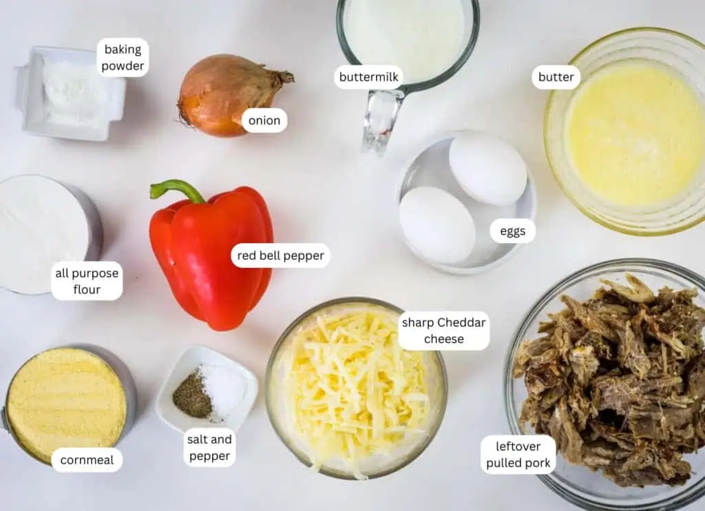 Ingredients for cornbread with pulled pork are arranged on a white surface, including flour, cornmeal, baking powder, onion, red bell pepper, salt, pepper, buttermilk, eggs, butter, cheese, and pulled pork.