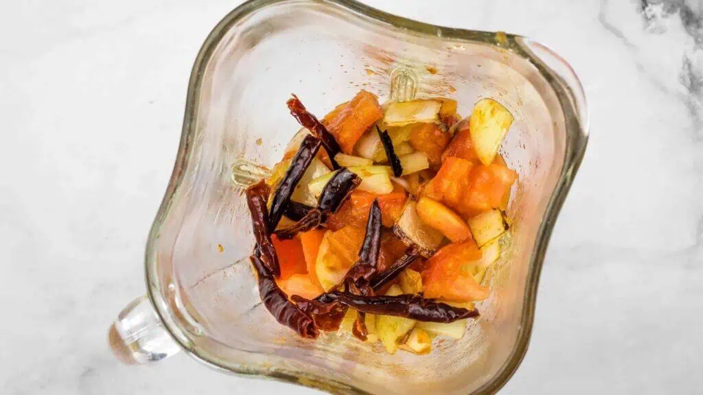 Chopped tomatoes, dried red chilies, garlic, and onion pieces inside a blender jar on a white surface.