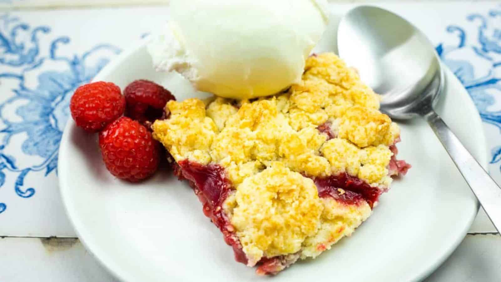 A slice of berry cobbler topped with a scoop of vanilla ice cream is served on a white plate with a spoon and fresh raspberries.