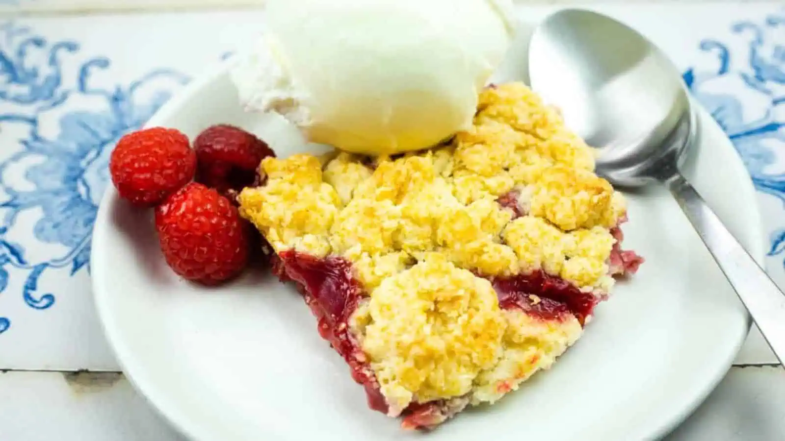 A slice of berry cobbler topped with a scoop of vanilla ice cream is served on a white plate with a spoon and fresh raspberries.
