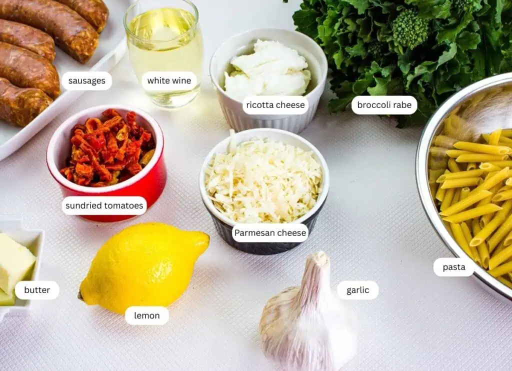 An assortment of labeled ingredients on a counter: sausages, white wine, ricotta cheese, broccoli rabe, pasta, garlic, lemon, butter, sundried tomatoes, and Parmesan cheese.