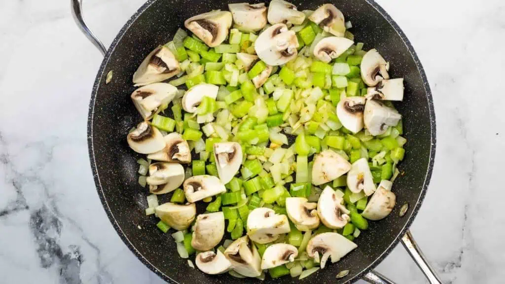 Chopped celery, onions, and quartered mushrooms cooking in a black skillet on a white marble surface.