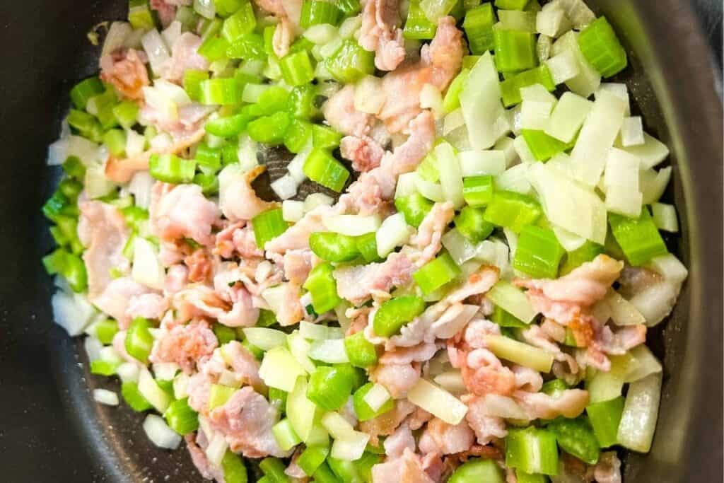 Chopped celery, onion, and pieces of bacon cooking together in a pan.
