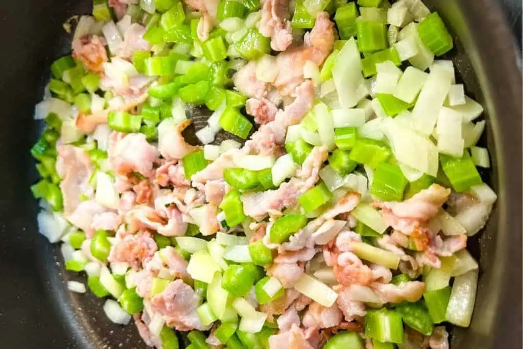 Chopped celery, onion, and pieces of bacon cooking together in a pan.
