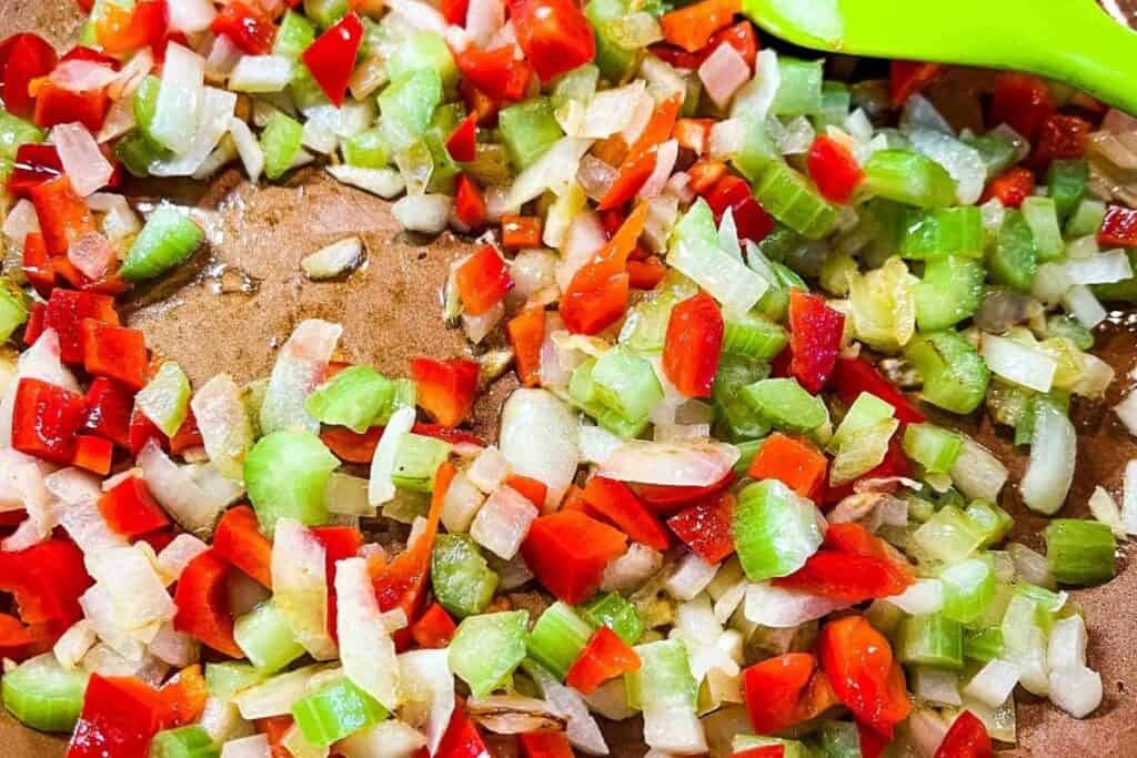 Chopped red bell peppers, onions, and celery being saut&eacute;ed in a pan with a green spatula.