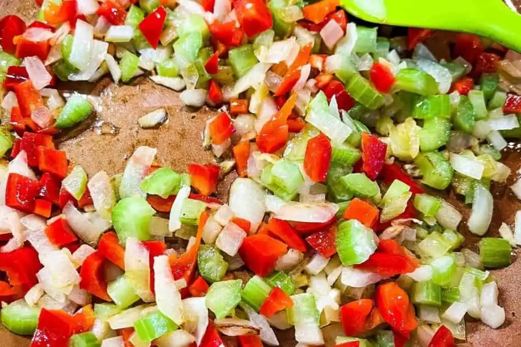Chopped red bell peppers, onions, and celery being sautéed in a pan with a green spatula.