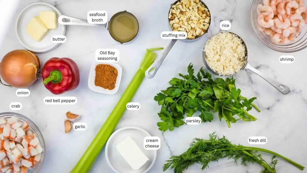 Assorted ingredients on a countertop, including onion, red bell pepper, garlic, crab, butter, seafood broth, Old Bay seasoning, celery, cream cheese, stuffing mix, rice, parsley, dill, and shrimp.