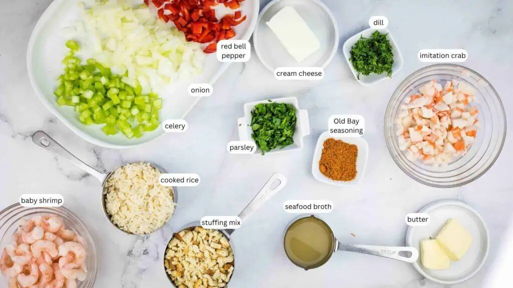 Ingredients for a seafood dish arranged on a counter, including chopped vegetables, herbs, seafood, stuffing mix, butter, cream cheese, Old Bay seasoning, and seafood broth.