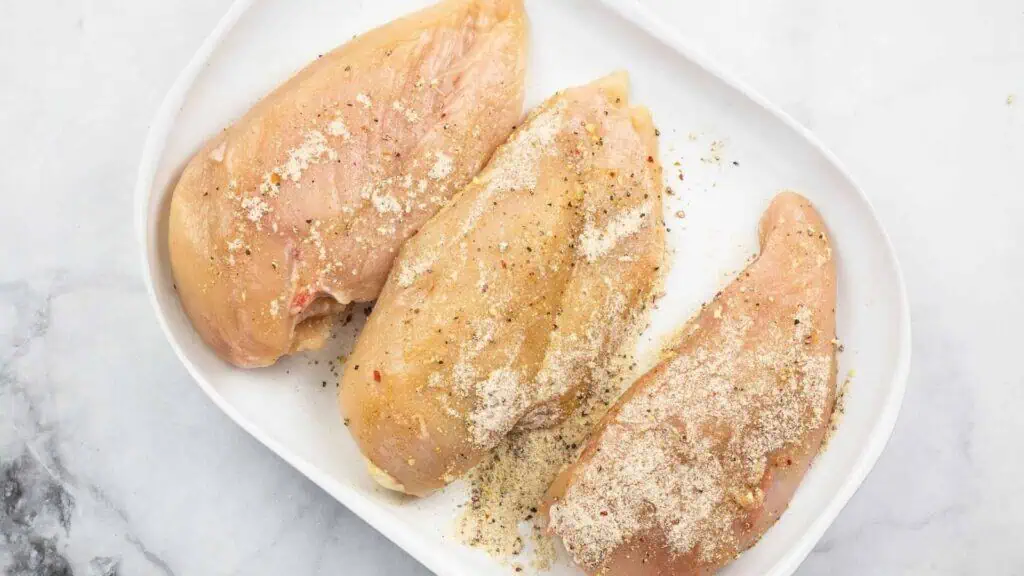 Three raw chicken breasts on a white plate, sprinkled with seasoning and black pepper, placed on a light marble surface.