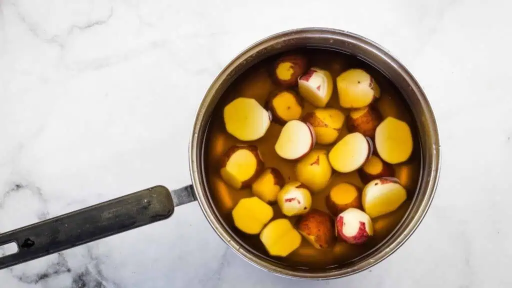 A saucepan filled with peeled and halved potatoes submerged in water, placed on a white marble surface.