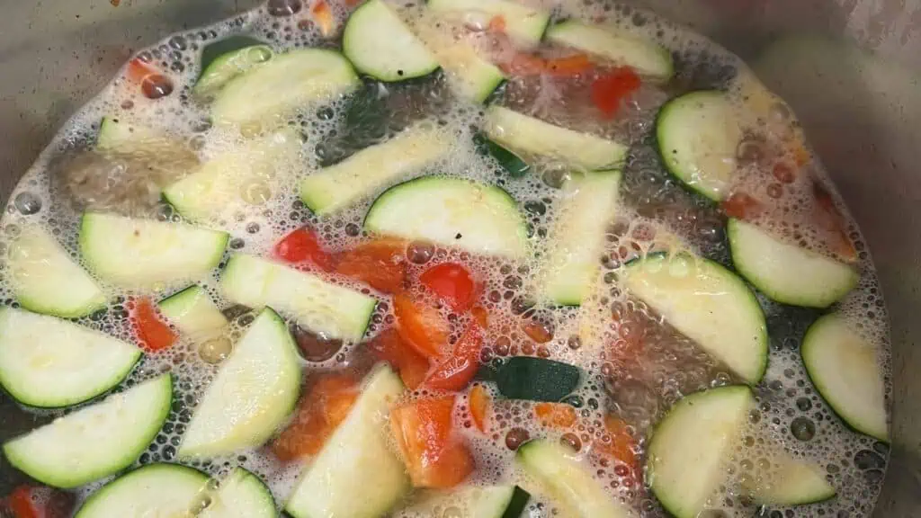 Sliced zucchini and red bell pepper cooking in boiling water inside a pot.