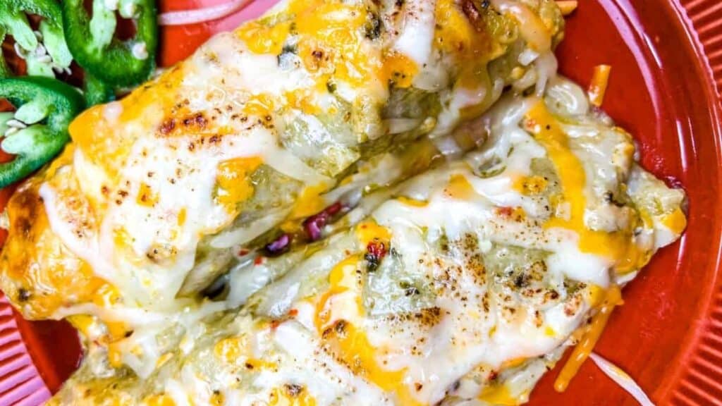 Two cheesy enchiladas topped with melted cheese are served on a red plate, with sliced jalapeños on the side.