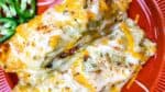 Two cheesy enchiladas topped with melted cheese are served on a red plate, with sliced jalapeños on the side.